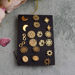 Könnte beinhalten: Ein Set mit 16 goldfarbenen Ohrringen mit verschiedenen himmlischen und geometrischen Designs, darunter Monde, Sonnen, Unendlichkeitszeichen und Lebensblumenmuster. Die Ohrringe werden auf einem schwarzen Hintergrund präsentiert.