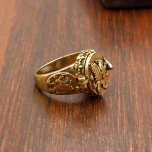 May include: A gold-colored ring with detailed relief carvings. The ring features a central circular emblem with a bird design and additional decorative elements on the band. The ring is set against a dark wooden surface.