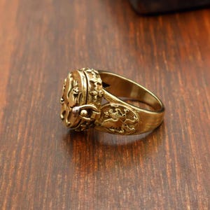May include: A gold-colored ring with an oval-shaped face and ornate detailing. The ring features a hinged compartment and decorative relief patterns, possibly depicting mythical creatures. The ring is set on a wooden surface.
