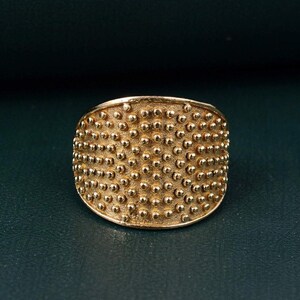 May include: A wide gold-toned ring featuring a textured surface covered in a dense, uniform pattern of small, raised spherical dots. The metal has a matte, slightly brushed finish, creating a consistent appearance across the entire band.
