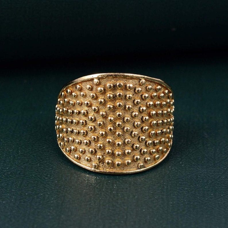 May include: A wide gold-toned ring featuring a textured surface covered in a dense, uniform pattern of small, raised spherical dots. The metal has a matte, slightly brushed finish, creating a consistent appearance across the entire band.