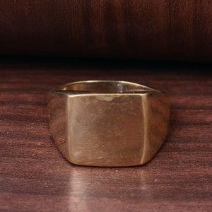 Op de afbeelding: Een gouden zegelring met een vierkante bovenkant. De ring is gemaakt van glad, glanzend metaal.