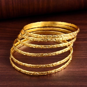 May include: A set of six gold-toned bangles with intricate engraved designs. The bangles are stacked on top of each other, creating a layered look.