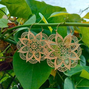 May include: A pair of gold-colored, filigree flower earrings. The earrings are hanging from a green branch with green leaves in the background. The earrings have a detailed, openwork design, with a floral pattern.