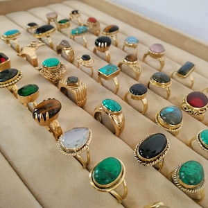 Könnte beinhalten: Eine Sammlung von Goldringen mit verschiedenen Edelsteineinsätzen. Die Ringe werden in einer Holzkiste mit einzelnen Fächern präsentiert. Die Edelsteine umfassen Türkis, Tigerauge, Labradorit, Smaragd und schwarzer Onyx.