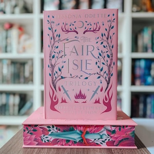 May include: A pink hardcover book titled The Fair Isle Trilogy Deluxe Ultimate Collection by Tessonja Odette, featuring illustrations of trees, a crown, and swords. The book rests on a matching pink box with a decorative sword and floral design.