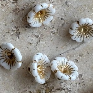 Puede incluir: Una colección de pequeñas cuentas con forma de flor que presentan un acabado blanco opaco con detalles en oro metálico que irradian desde el orificio central. Cada cuenta tiene un borde festoneado y una estructura cóncava de pétalos.