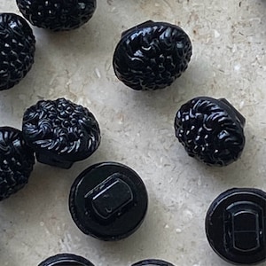 Puede incluir: Una colección de botones negros redondos con un patrón floral texturizado y elevado en la parte frontal y una parte posterior lisa y plana con un vástago sobresaliente para su fijación. Los botones tienen un acabado brillante y disperso.