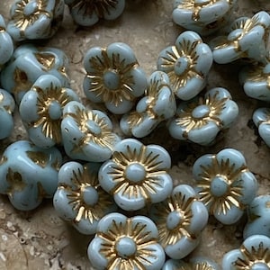 Op de afbeelding: Een verzameling kleine, lichtblauwe bloemvormige kralen met vijf bloemblaadjes. Elke kraal heeft een verhoogd, metallic goudkleurig midden en goudkleurige details die vanuit het midden van de bloem naar de randen van elk bloemblaadje stralen.