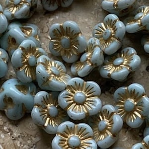 Puede incluir: Un conjunto de cuentas en forma de flor de color azul claro con centros dorados. Las cuentas tienen un acabado brillante y están dispersas sobre una superficie. Ideales para la fabricación de joyas y proyectos de artesanía.