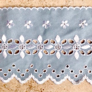 Może przedstawiać: Biała koronka bawełniana z falowanym brzegiem i delikatnym wzorem kwiatowym. Koronka jest na jasnoniebieskim tle.