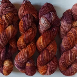 Könnte beinhalten: Vier Stränge handgefärbter Wolle in einem Farbverlauf warmer Farben. Das Garn wechselt von tiefem Burgunder zu leuchtenden Orange- und Gelbtönen. Die Stränge sind eng gewickelt und zeigen die Farbvariationen.