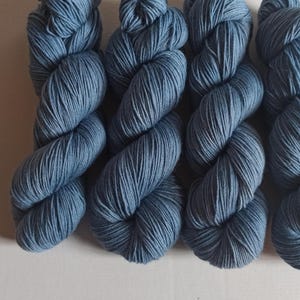 Könnte beinhalten: Vier Stränge blaue Wolle sind zu sehen. Die Wolle hat eine einfarbige, mittelblaue Farbe. Die Wolle ist eng zu einer verdrehten, seilartigen Form gewickelt, mit sichtbaren Strähnen. Der Hintergrund ist eine schlichte, cremefarbene Oberfläche.