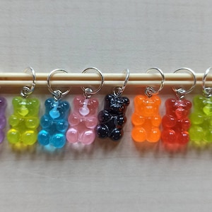 Könnte beinhalten: Ein Set aus zehn bunten Gummibärchen-Maschenmarkierern, jeder mit einem silbernen Ring. Die Marker sind in einer Reihe auf einer Holzoberfläche angeordnet.