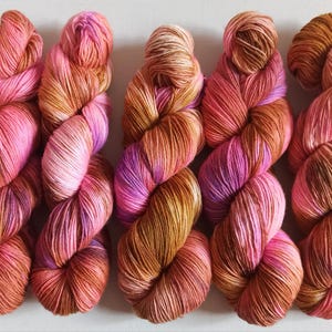 Könnte beinhalten: Fünf handgefärbte Garnstränge, die nebeneinander angeordnet sind. Jeder Strang weist eine melierte Farbpalette aus leuchtendem Pink, sanftem Magenta, warmem Kupfer und tiefen goldbraunen Tönen auf, die zu einem traditionellen Strang gedreht sind.