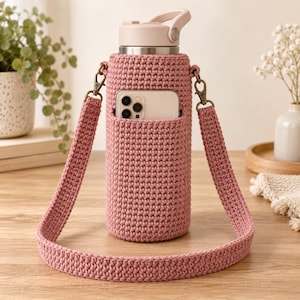 May include: A dusty rose crochet water bottle holder featuring a matching adjustable shoulder strap and a front pocket containing a smartphone. The holder is made of textured yarn and includes bronze-colored metal swivel clips for the strap.