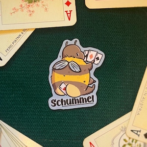 Könnte beinhalten: Ein Aufkleber mit einer cartoonartigen, bienenähnlichen Figur, die Spielkarten hält, mit dem Wort "Schummel" darunter. Der Aufkleber ist von Vintage-Spielkarten mit floralen Designs und dem Ass jeder Farbe umgeben.