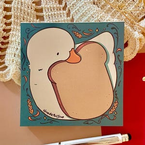 Op de afbeelding: Een vierkant turquoise notitieblok met een illustratie van een gans die een sneetje toast omhelst. Het notitieblok heeft een decoratieve rand en de tekst "Nevada Art Shop". Een witte styluspen ligt eronder.