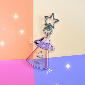 May include: A silver keychain with a star charm and a clear plastic charm featuring a purple UFO with a white bird inside. The charm is filled with glitter.