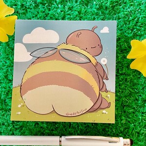 Pode incluir: Uma impressão de arte quadrada com uma abelha de desenho animado com abdómen listrado, asas e uma cabeça pequena. O fundo mostra um céu azul com nuvens e um campo gramado. A impressão está rotulada como "Nevada Art Shop".