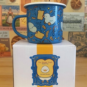 Könnte beinhalten: Blaue Emaille-Tasse mit Enten-, Brot- und Weizen-Motiv. Die Tasse steht auf einer weißen Schachtel mit der Aufschrift "The Bread Thief Enamel Cup" und einer Enten-Illustration.