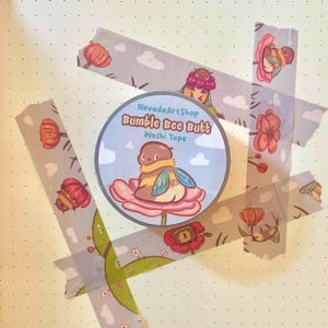 Può includere: Un rotolo di nastro washi con un design a fumetto di un sedere di ape, circondato da strisce di nastro washi con motivi floreali e di api. Il rotolo di nastro ha il testo "Bumble Bee Butt" e "NevadaArtShop". Lo sfondo è una carta a pois giallo chiaro.