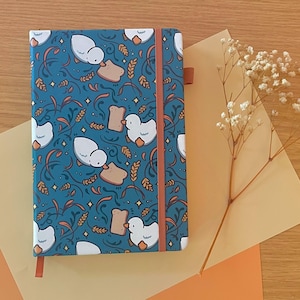 Peut inclure: Un carnet de notes bleu sarcelle avec un motif fantaisiste de canards blancs, de pain grillé et d'épis de blé. Il est doté d'une bande élastique orange et d'un signet en ruban assorti. Un accessoire de papeterie.