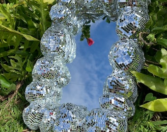 May include: A rectangular mirror with a frame composed of many small, reflective disco ball pieces. The mirror reflects a blue sky with clouds. The frame is silver and the mirror is surrounded by green foliage and grass.
