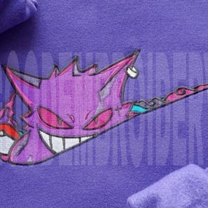 May include: Purple hoodie featuring an embroidered design of a purple cartoon character with red eyes and a wide grin. The character is incorporated into a swoosh design, holding a red and white ball. The hoodie is made of a soft, comfortable material.