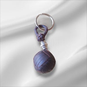 Può includere: Un portachiavi in pelle marrone scuro con un design a sfera intrecciata. Il portachiavi presenta un anello in metallo color argento e un accento decorativo in metallo color argento. La sfera ha un diametro di circa 5 cm.