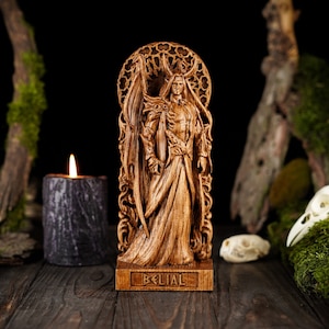 Könnte beinhalten: Eine Holzstatue einer geflügelten Figur, die in einem geschnitzten Rahmen steht. Die Statue ist aus dunklem Holz gefertigt und weist detailreiche Verzierungen auf. Der Sockel der Statue ist mit dem Wort "Belial" beschriftet.