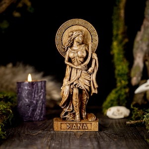 May include: Wooden statue of the Roman goddess Diana, standing with a bow and arrow, with a Greek key design around her head. The statue is on a wooden base with the word "DIANA" inscribed on it.
