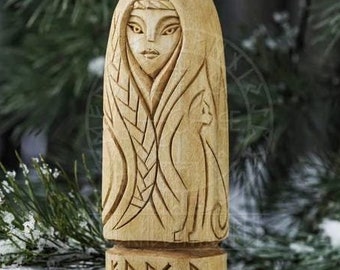 May include: A wooden carving of a woman with long hair and a cat. The carving is in a natural wood color and has intricate details. The woman is wearing a long robe and has a serene expression on her face. The carving is sitting on a tree stump in a snowy forest.