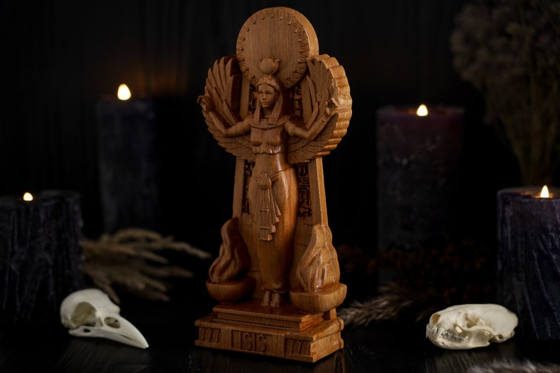 May include: A wooden statue of the Egyptian goddess Isis, standing on a base with the word "Isis" inscribed. The statue is surrounded by candles and skulls, creating a dark and mystical atmosphere.