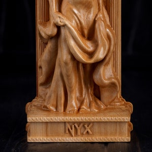 Puede incluir: Una escultura de madera de una mujer con una t&uacute;nica fluida, de pie sobre una base con la palabra "NYX" inscrita en ella.
