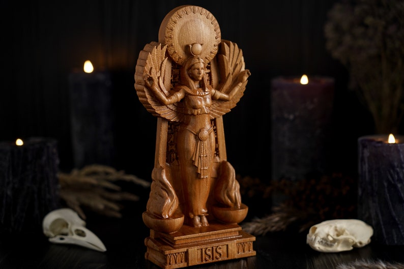 May include: A wooden statue of the Egyptian goddess Isis, standing on a base with the word "ISIS" inscribed. The statue is intricately carved with details of her wings, headdress, and clothing.