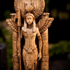 May include: A wooden statue of a winged goddess with a crown and a spear. The statue is carved in a detailed style and has a warm brown color.