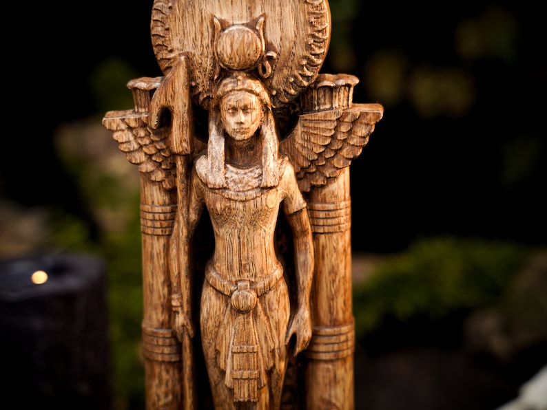 May include: A wooden statue of a winged goddess with a crown and a spear. The statue is carved in a detailed style and has a warm brown color.