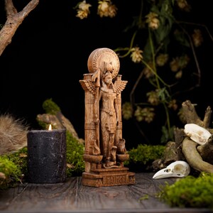 May include: A wooden statue of a goddess with wings, standing on a pedestal with the words "IN MAAT" inscribed. The statue is surrounded by moss, a candle, and a skull.