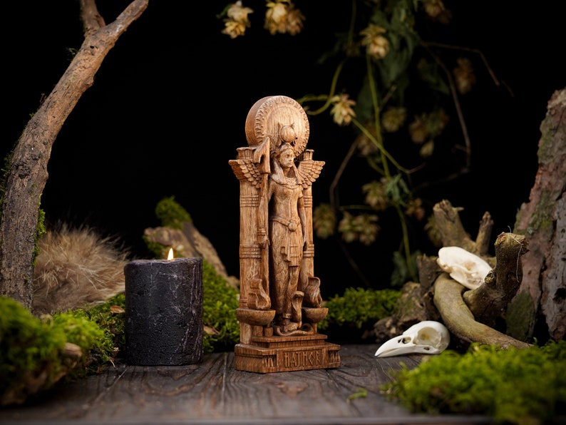 May include: A wooden statue of a goddess with wings, standing on a pedestal with the words "IN MAAT" inscribed. The statue is surrounded by moss, a candle, and a skull.