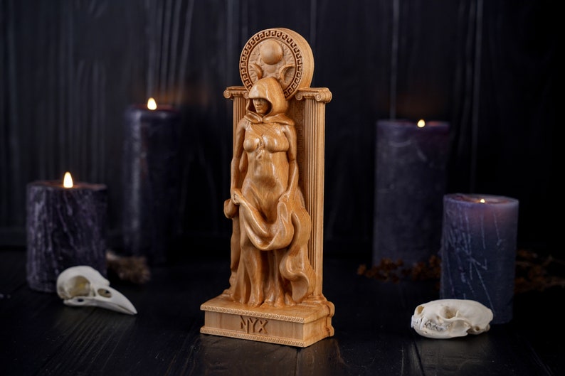 Puede incluir: Una estatua de madera de la diosa griega Nyx, representada como una mujer con capucha y t&uacute;nicas fluidas, de pie sobre un pedestal con la palabra "NYX" inscrita en &eacute;l. La estatua est&aacute; rodeada de velas negras y cr&aacute;neos.