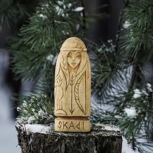 Puede incluir: Una talla de madera de una mujer con el pelo largo y una capucha, sosteniendo un arco y una flecha. La talla est&aacute; etiquetada como "Skadi".