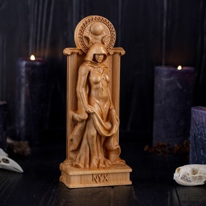 Puede incluir: Una estatua de madera de una figura femenina con capucha, de pie en una puerta con una luna creciente y el sol sobre su cabeza. La estatua est&aacute; etiquetada como "NYX" en la base.