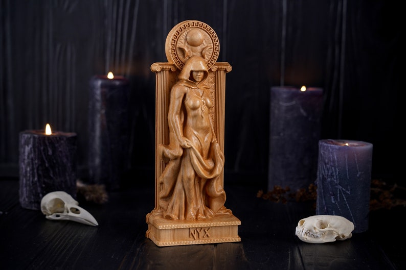 Puede incluir: Una estatua de madera de una figura femenina con capucha, de pie en una puerta con una luna creciente y el sol sobre su cabeza. La estatua est&aacute; etiquetada como "NYX" en la base.