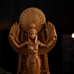 May include: A wooden statue of an Egyptian goddess with wings outstretched, wearing a crown and a headdress. The statue is standing on a pedestal with hieroglyphics carved into it. The statue is lit by candlelight.