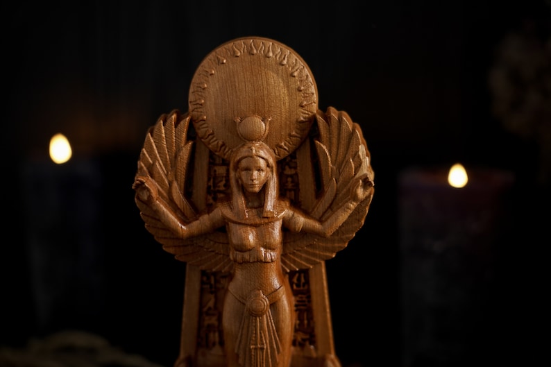May include: A wooden statue of an Egyptian goddess with wings outstretched, wearing a crown and a headdress. The statue is standing on a pedestal with hieroglyphics carved into it. The statue is lit by candlelight.
