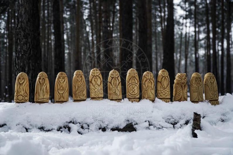Puede incluir: Una fila de 12 figuras de madera talladas con s&iacute;mbolos de la mitolog&iacute;a n&oacute;rdica, de pie sobre una superficie nevada en un entorno boscoso.