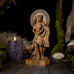 May include: A wooden statue of the Roman goddess Diana, carved in detail. She is depicted standing with a bow and arrow, and her name is inscribed on the base. The statue is set against a dark background with moss and a candle.