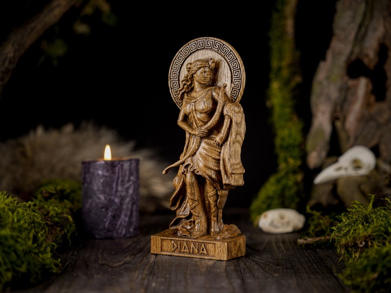 May include: A wooden statue of the Roman goddess Diana, carved in detail. She is depicted standing with a bow and arrow, and her name is inscribed on the base. The statue is set against a dark background with moss and a candle.
