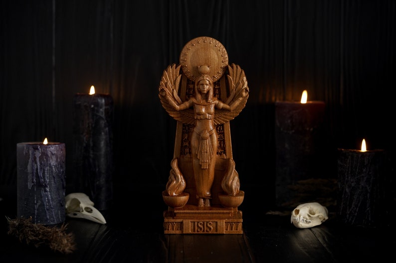 May include: A wooden statue of the Egyptian goddess Isis, standing on a base with the word "ISIS" inscribed. The statue is surrounded by black candles and white animal skulls.
